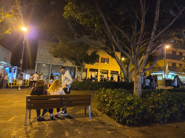 People sitting on a bench and standing under large trees in a square in Güímar while waiting for the night race to start.