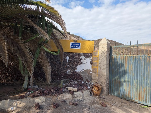 Old wall with a street sign reading Carretera Palm-Mar at the edge of the settlement with volcanic hills behind.