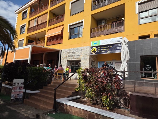 Café terrace beneath a yellow apartment building in Palm-Mar with people dining and a sign for All Properties & All Insurance Tenerife.