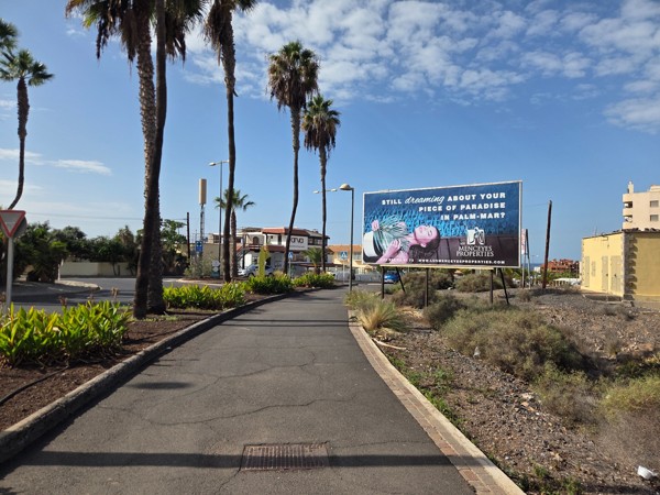 Landscaped promenade in Palm-Mar with palm trees, shrubs and the curved façade of a modern apartment building.