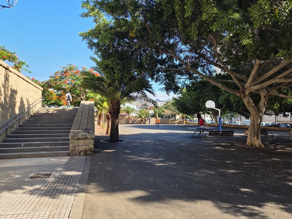Wide view of Parque El Quijote showing the outdoor basketball court, benches under large trees and open paved space.