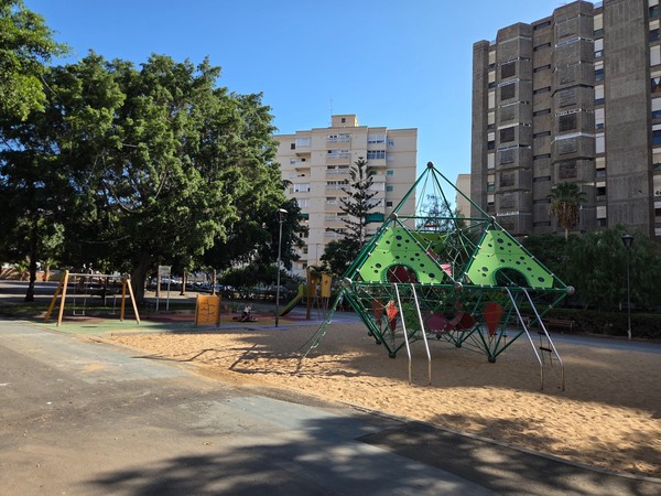 Modern children’s playground with large green rope-climbing pyramids set in a sand area, surrounded by apartment buildings and trees.