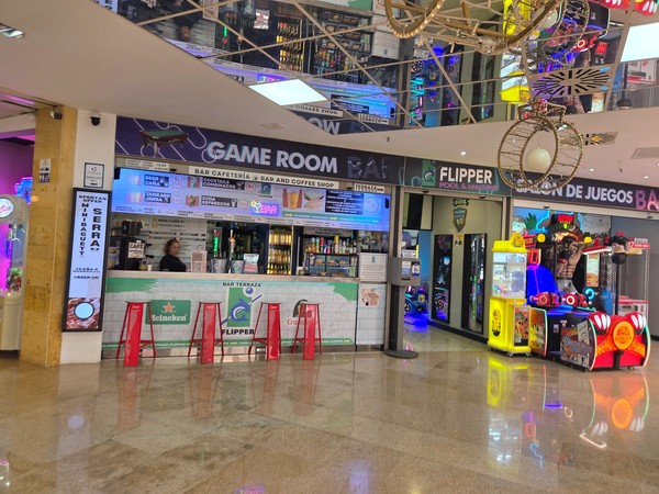 Flipper bar and coffee shop inside Pasarela Shopping Center with a long counter, illuminated drinks displays and arcade machines next to the seating area.