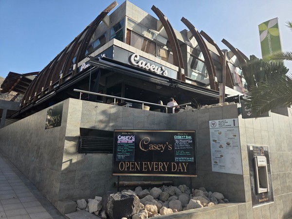 Exterior of Casey’s bar and restaurant on the upper level of Pasarela Shopping Center with modern stone walls, curved wooden beams and a sign that reads open every day.