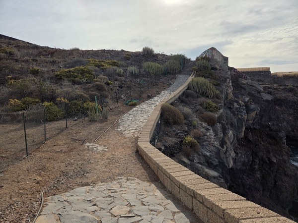 Narrow cliff-edge section of the paved path at Playa Aguadulce, lined with cacti and low volcanic vegetation.