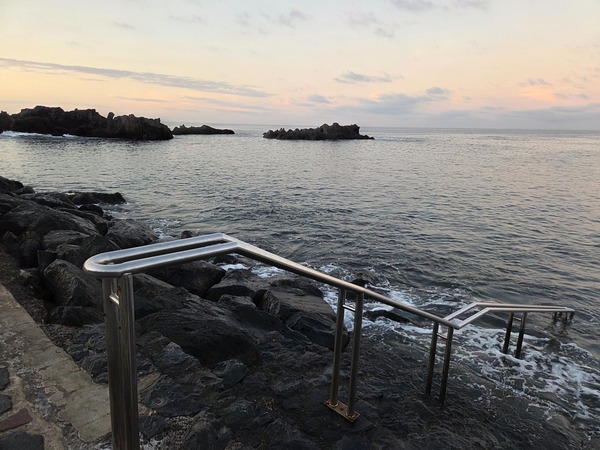 Stainless steel handrail leading into the water over volcanic rocks for safe sea entry.