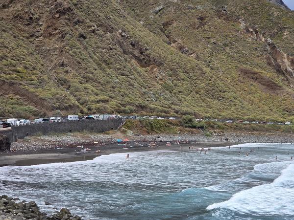 Panoramic view of Playa de Almáciga with black sand beach, swimmers, and a backdrop of green mountains.