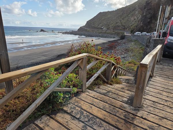Wooden stairs leading down to Playa de Almáciga, bordered by plants and volcanic rocks.