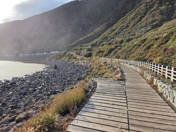 Wooden boardwalk along Playa de Almáciga with volcanic rocks and lush hillsides.