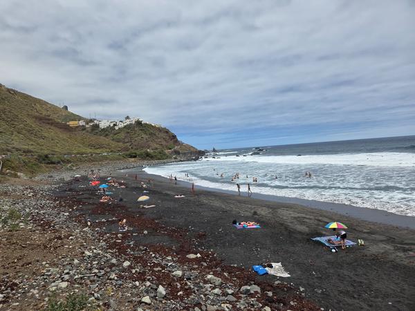 People enjoying the surf and sunbathing on Playa de Almáciga under a cloudy sky.