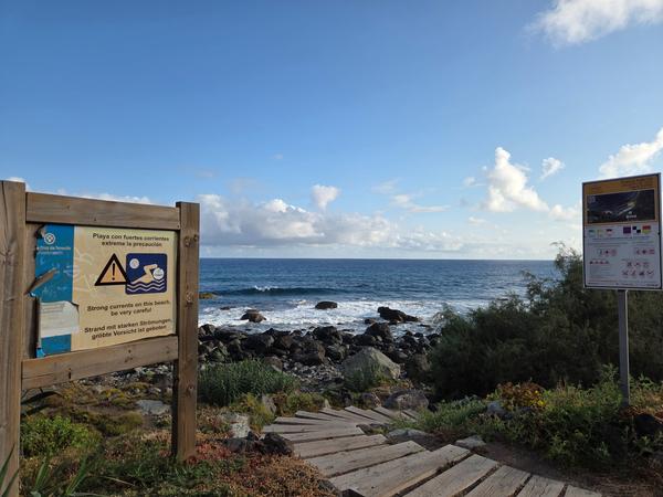 Warning signs about strong currents at Playa de Almáciga in Spanish, English, and German.