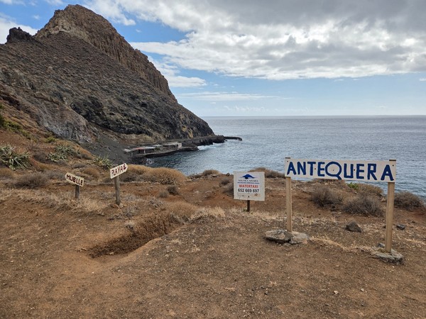 Wooden signs on the cliff edge marking Playa de Antequera, with arrows pointing to the pier and the beach above the Atlantic coast.