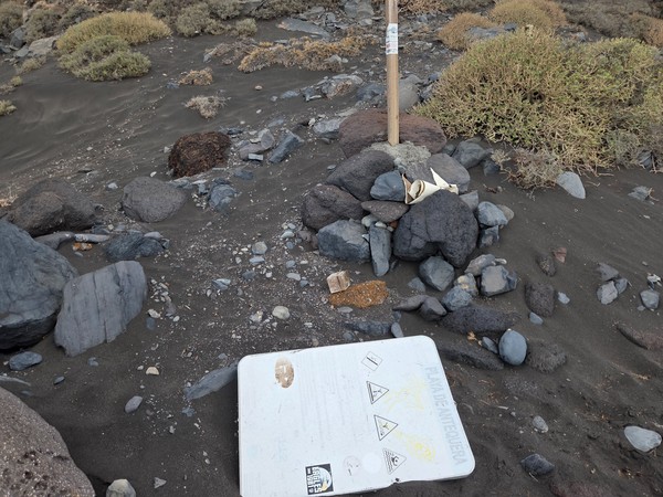 Broken information sign for Playa de Antequera lying on the dark sand among volcanic rocks and dry coastal plants.