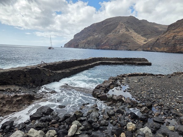 Stone pier and natural pool area at Playa de Antequera with a catamaran anchored offshore and steep volcanic cliffs in the background.