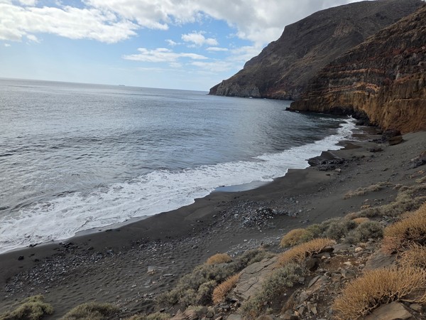 Low Atlantic waves breaking on the black sand of Playa de Antequera with a rocky headland in the background.