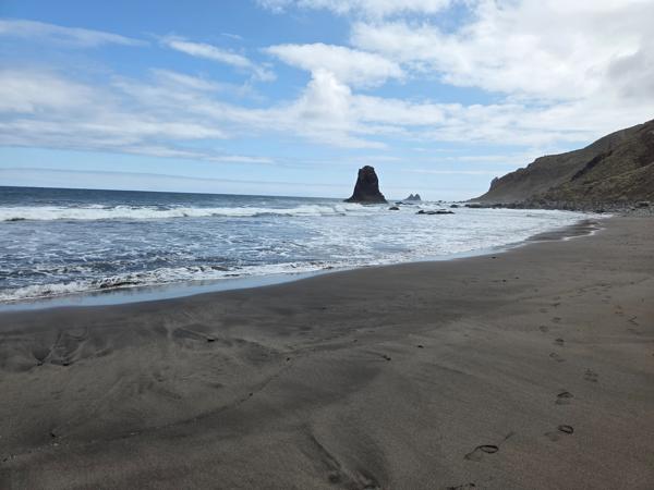 Wide black sand shore of Playa de Benijo with visible footprints and rugged rock in the sea.