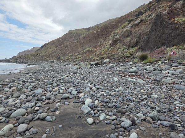 Rocky section of Playa de Benijo with large rounded stones and a cliff warning sign on the slope.