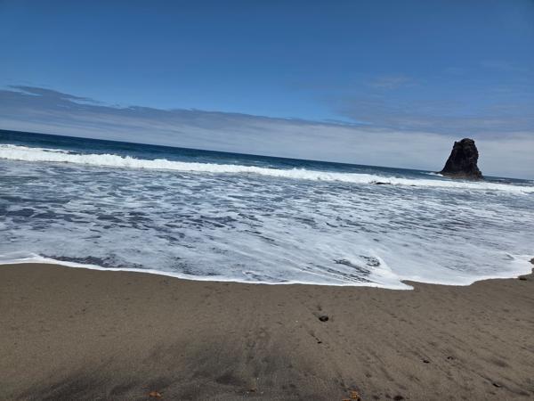 Foamy waves rolling onto Playa de Benijo’s volcanic sand, with the tall sea stack under a blue sky.