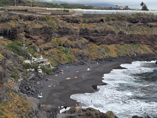 Zoomed-in cliffside view showing sunbathers scattered across the dark sand of the secluded cove.
