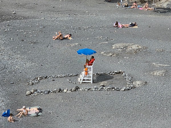 Lifeguard seated under a blue umbrella on Playa de Ajabo, surrounded by marked stones on the black sand.