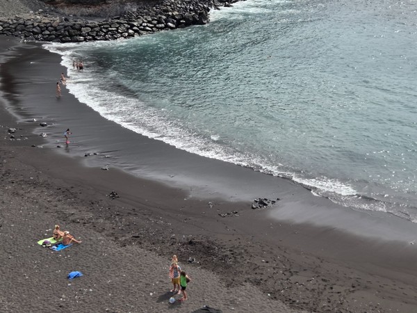Diagonal view of Playa de Ajabo’s shoreline with fine black sand, scattered pebbles and gentle surf.