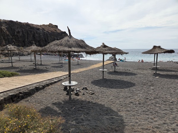 Row of straw parasols with round side tables on Playa de Ajabo, with the sea and cliffs in the background.