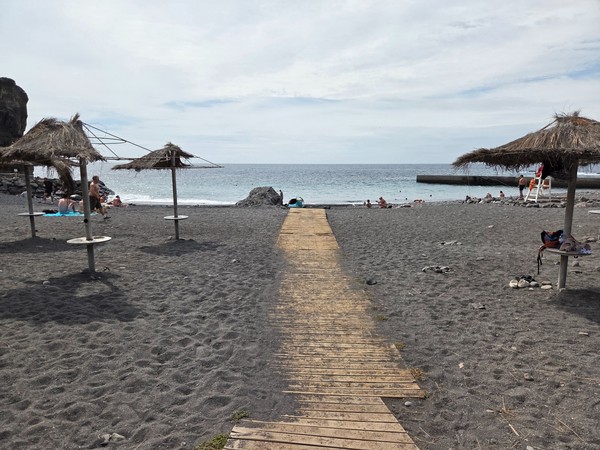 Wooden path running through the black sand towards the sea at Playa de Ajabo, passing between parasols.