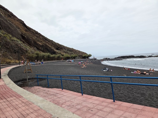 Paved access ramp with a blue handrail leading down to the black sand of Playa de la Arena in Tacoronte, Tenerife.