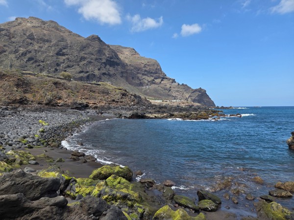 Crescent-shaped cove of Playa de Las Arenas framed by cliffs and emerald mossy rocks.