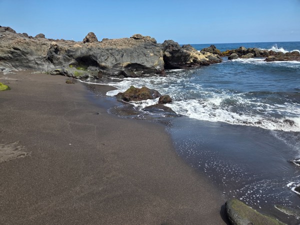 Pocket of fine black sand washed by gentle waves beside volcanic outcrops.