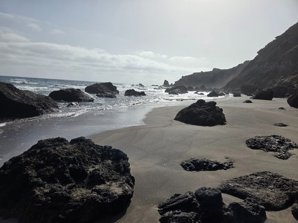 Close-up of Playa de las Breñas black sand with scattered lava boulders and foamy Atlantic surf.
