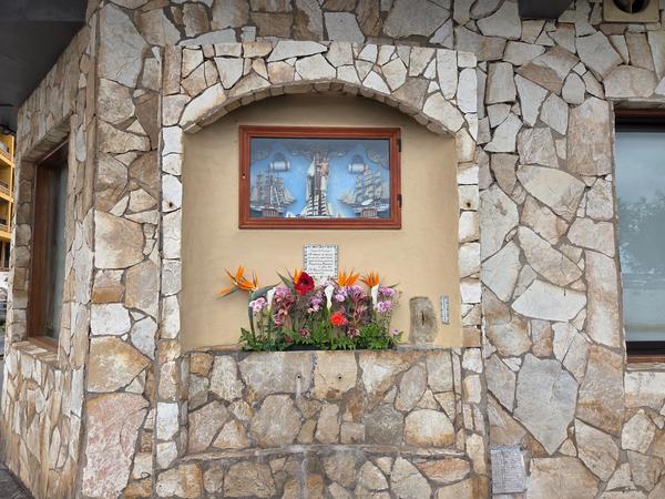 Small devotional shrine embedded in a stone wall with flowers and an image of the Virgin and ships.