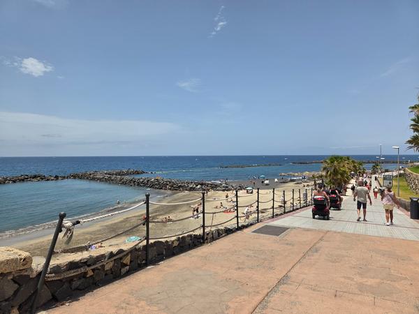 Oceanfront walkway beside Playa del Duque with palm trees and resting areas.