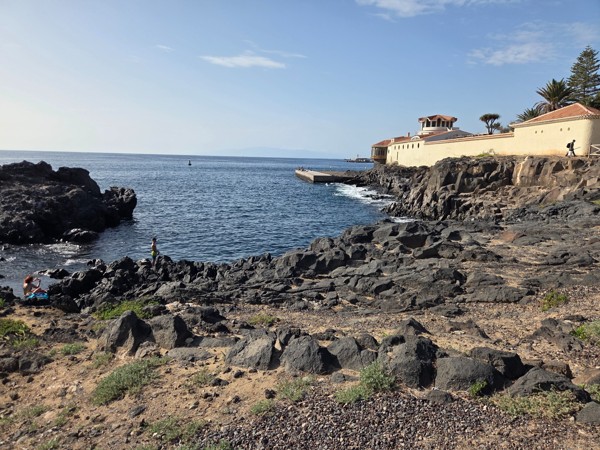 Volcanic rocks and a small natural inlet at Playa El Callao, with swimmers enjoying the calm Atlantic water next to the coastal path and villas.