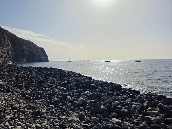 Pebble beach and dark cliff at Playa El Callao with several sailing boats and catamarans anchored in the calm, sunlit sea.