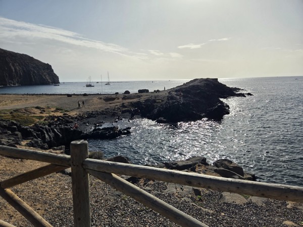 View from the wooden fence across a small rocky cove at Playa El Callao with people sitting near the water and yachts anchored offshore.