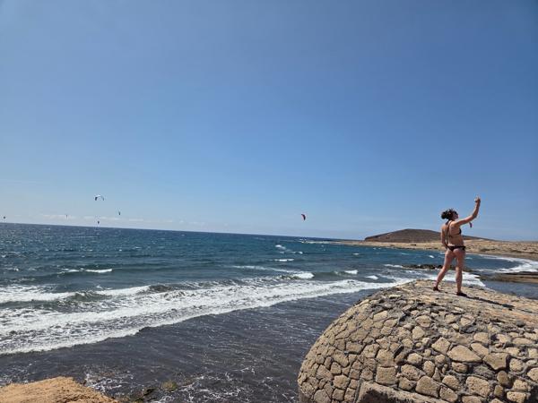 Woman taking a selfie on a stone platform with kitesurfers in the background at Playa El Médano.