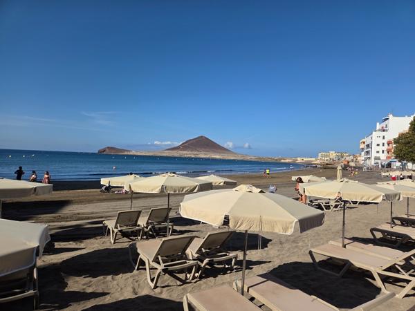 Rows of sunbeds and umbrellas on Playa El Médano with Montaña Roja in the background.
