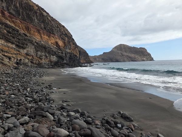 View along Playa Itamar Diez from the shore, with rounded volcanic stones in the foreground and smooth black sand curving towards the rocky point.