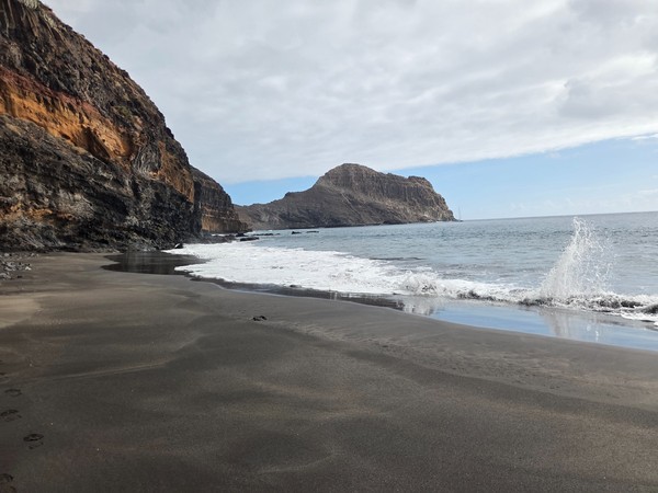 Foamy Atlantic wave breaking on the empty black sand of Playa Itamar Diez with the cliff face and headland in the background.