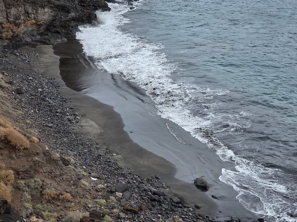 High-angle view of Playa Itamar Diez showing the narrow strip of wet black sand, scattered boulders and the breaking surf of the Atlantic Ocean.