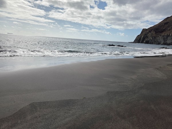 Black sand at Playa Itamar Diez with footprints leading towards the cliffs and the sea reflecting the cloudy sky.