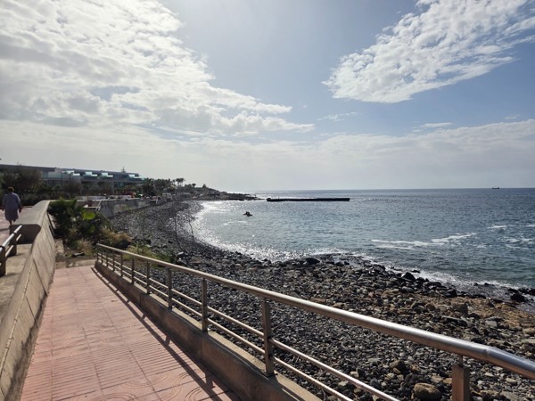 Walkway and railing leading down to the rocky shoreline of Playa La Arenita with the sea and breakwater in the background.