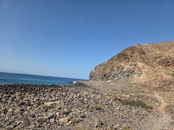 Volcanic cliff and coastal path extending from Playa La Arenita towards a rugged headland under a clear blue sky.