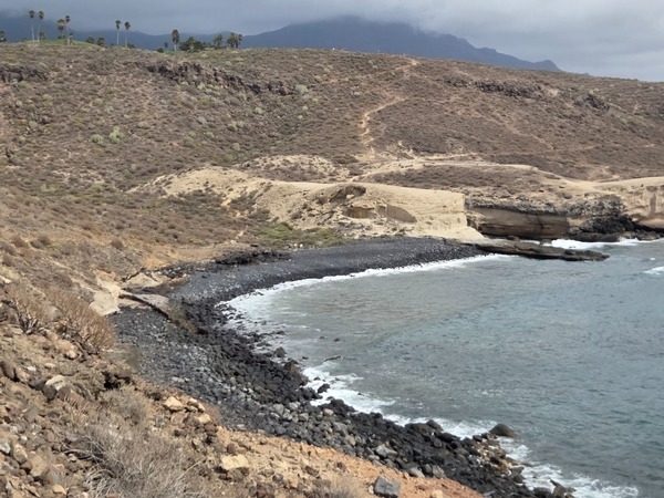 Wide view of Playa Pardela showing the curved pebble cove beneath arid hills and footpaths.