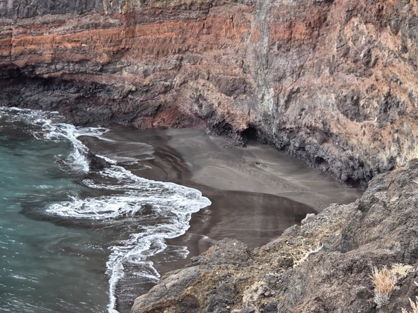 Closer view of the hidden black sand cove at Playa de Zapata, framed by dark volcanic cliffs and a small sea cave at the base of the rock wall.