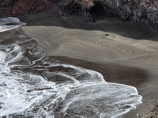Detailed close-up of the black sand at Playa de Zapata where gentle waves leave white foam patterns on the dark shoreline beneath the cliffs.