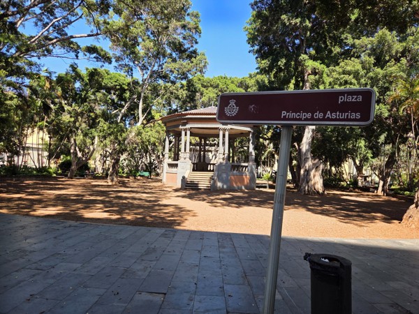 Street sign reading 'plaza Príncipe de Asturias' set against the trees and bandstand.