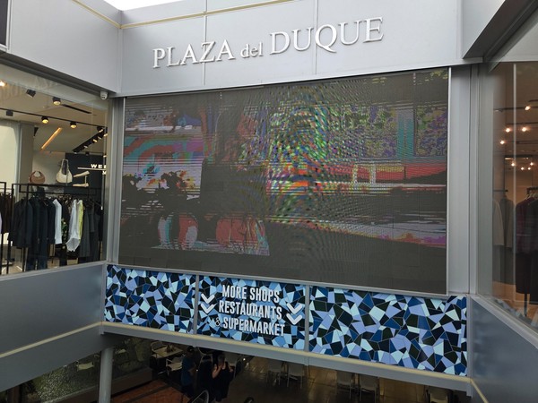 Large LED screen and Plaza del Duque sign at the central junction between levels.