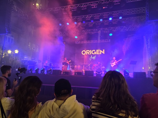 Concert scene with colored stage lights and the word Origen on the screen during a Plenilunio performance in Santa Cruz.
